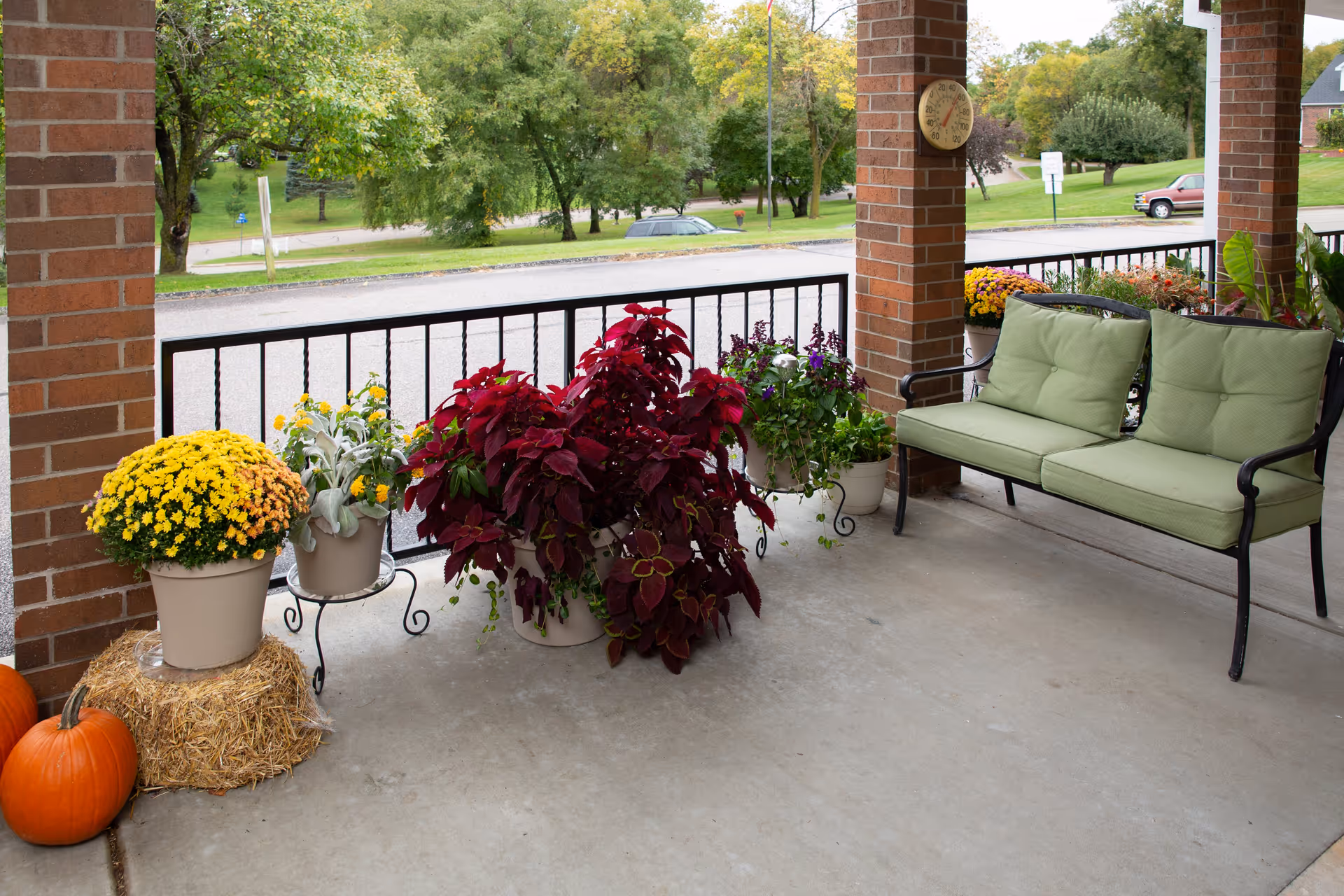 A covered outdoor patio area with a green cushioned bench, several potted plants with colorful flowers, two pumpkins on the ground, and a hay bale. The patio has brick pillars and overlooks a street with trees and parked cars.