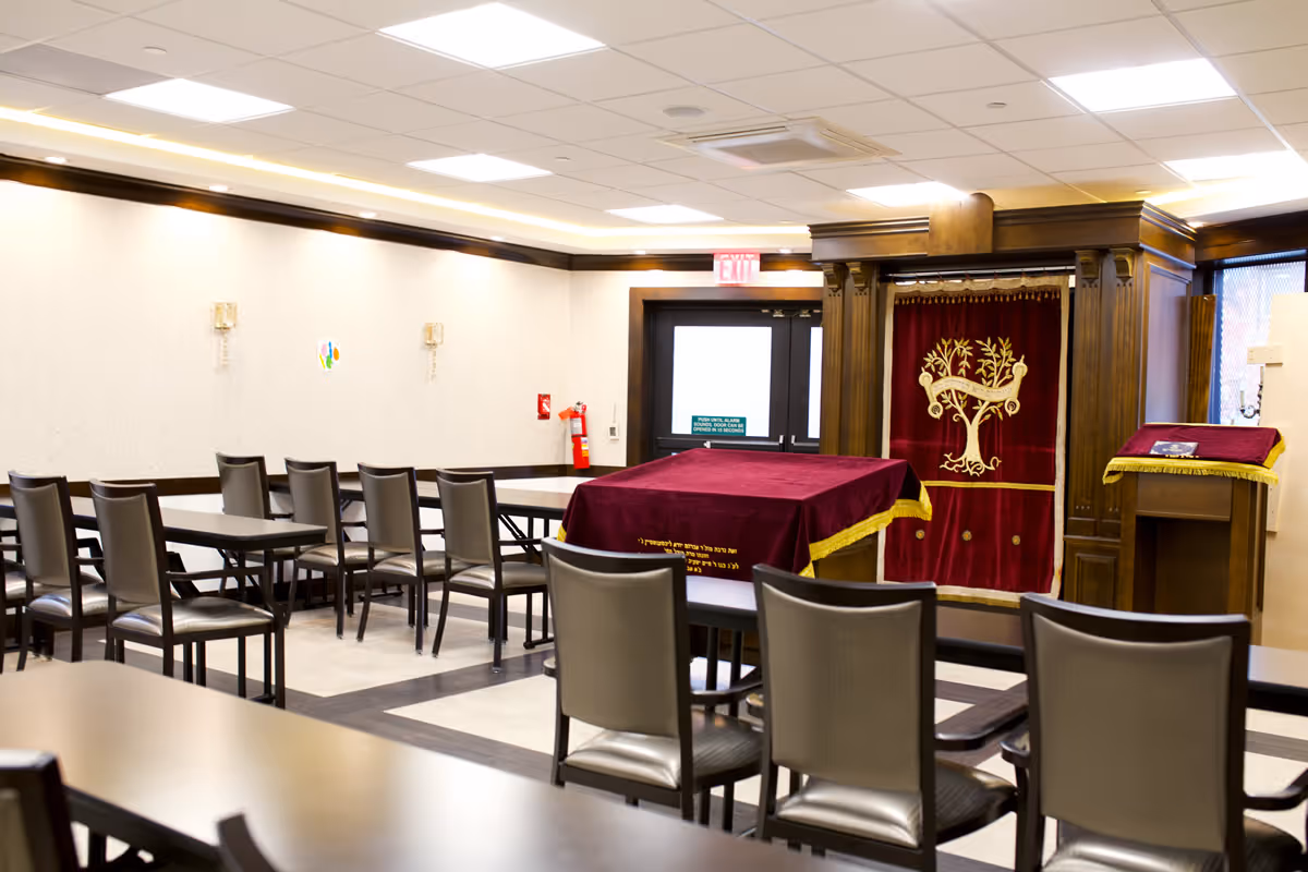 Interior view of a room with multiple tables and chairs arranged in rows, featuring a wooden cabinet with a red velvet curtain embroidered with a gold tree design, and a lectern covered with a matching red velvet cloth with gold trim.