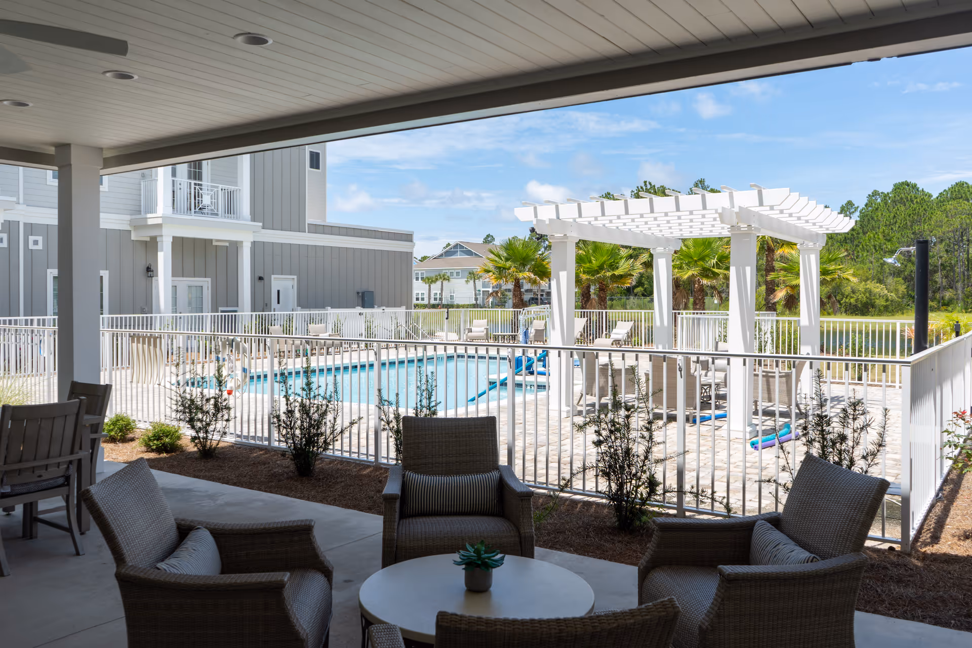 Covered outdoor seating area with wicker chairs and a round table overlooking a fenced swimming pool and a white pergola. The background includes palm trees, a gray building, and a clear blue sky.