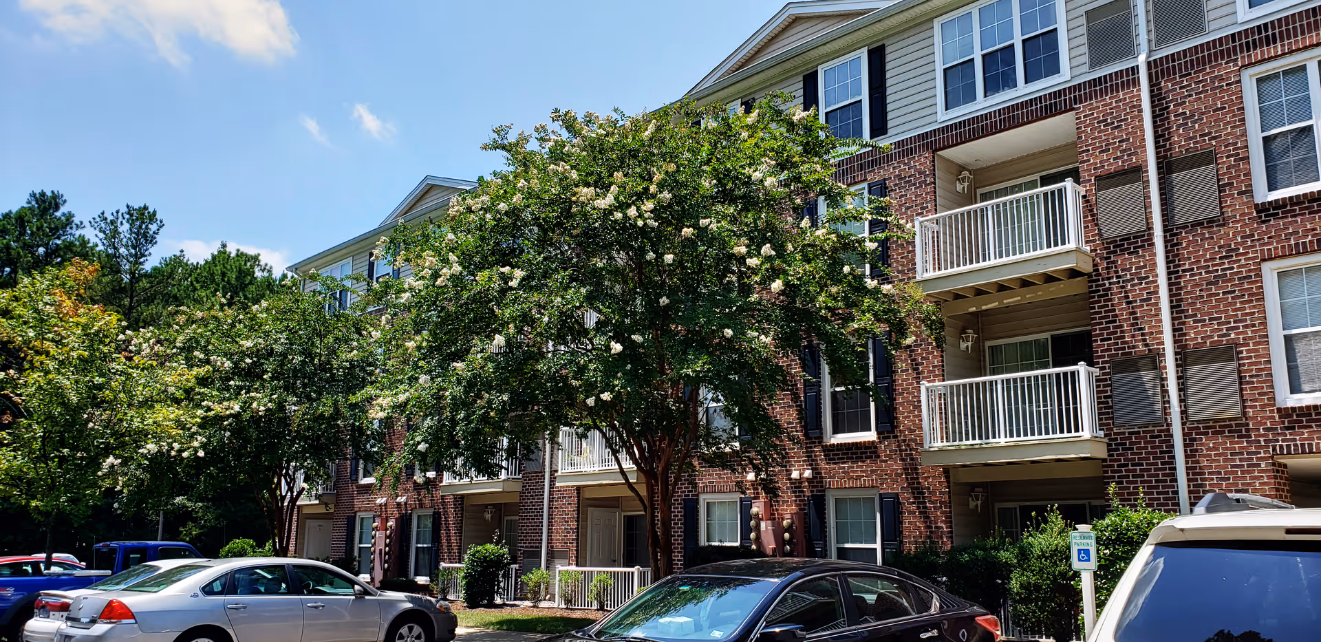 Brick three-story apartment building with balconies, trees, and parked cars in front under a blue sky.