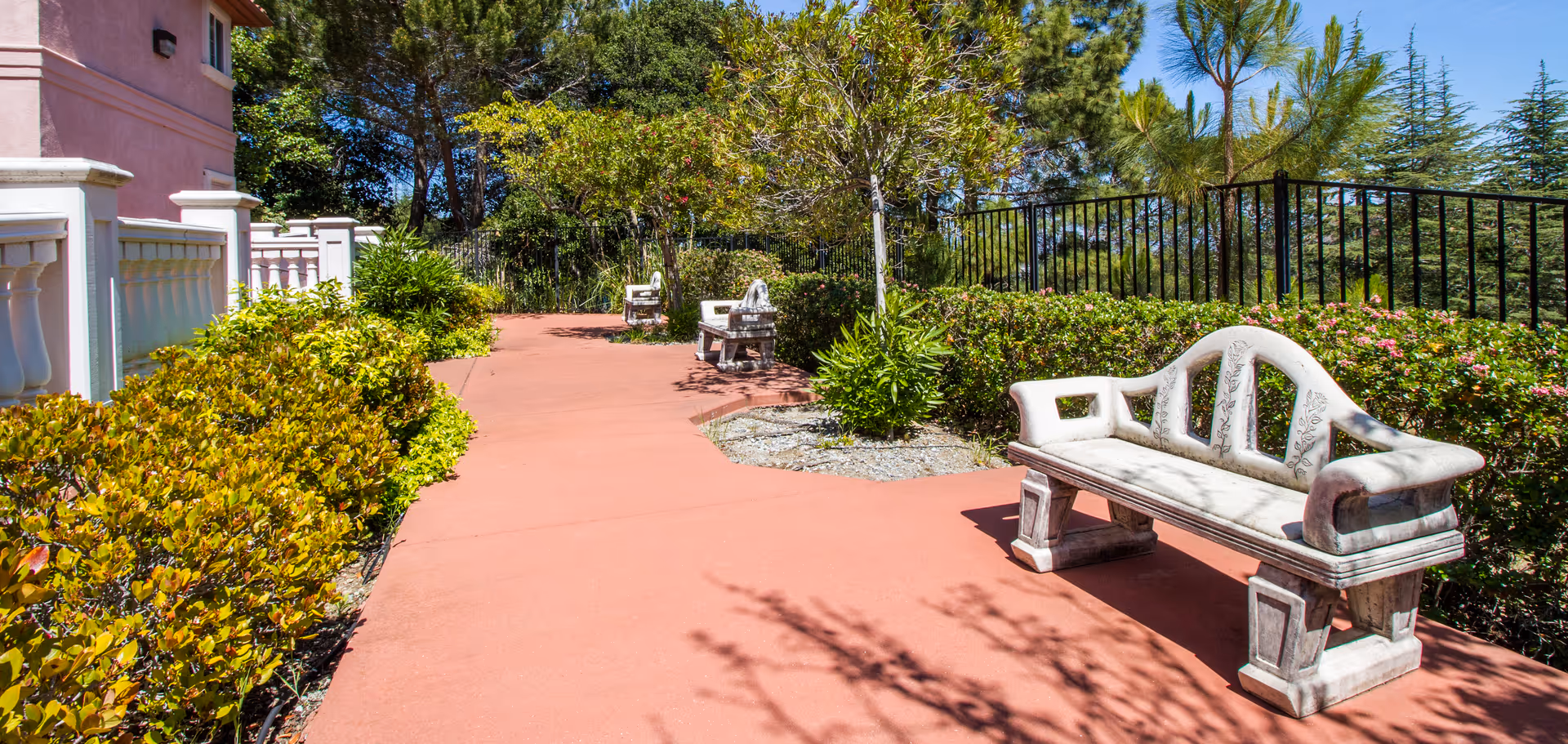 Outdoor garden pathway at Bayview Villa with red pavement, white stone benches, green bushes, trees, and a black metal fence under a clear blue sky.