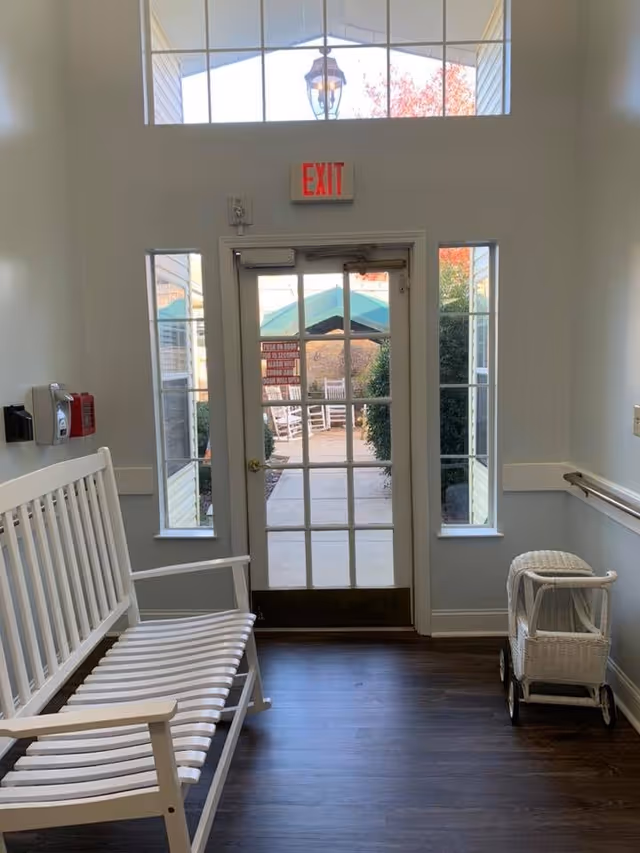 Interior view of a hallway in a senior living facility with a white wooden bench on the left and a small white wicker baby carriage on the right. A glass door with multiple panes leads outside to a patio area with rocking chairs and greenery. Above the door is a red EXIT sign and a large window letting in natural light.
