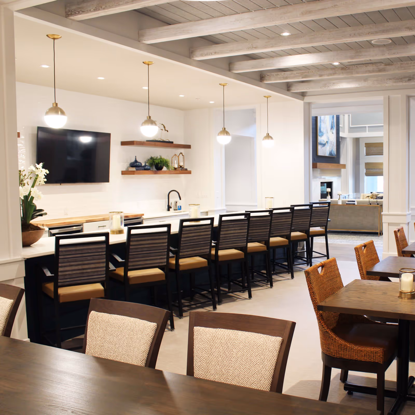 A modern dining area in a senior living facility featuring a long counter with eight high chairs, pendant lights hanging above, a wall-mounted TV, and shelves with decorative items. In the foreground, there are wooden tables with cushioned chairs, and in the background, a living room area with a fireplace and large windows is visible.
