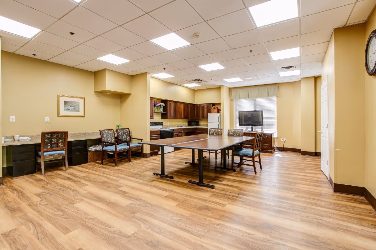 A spacious room with wooden flooring featuring a kitchen area with dark wood cabinets, a white refrigerator, and a stove. In the center, there is a table tennis table surrounded by chairs. The room has beige walls, a large window with blinds, a wall clock, and a TV on a stand near the window.