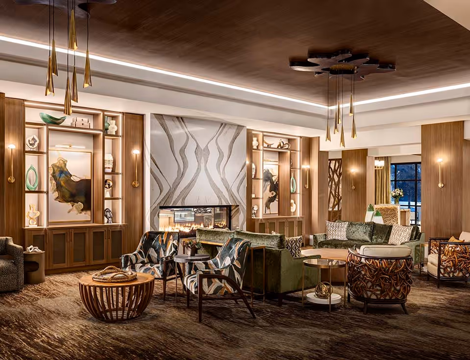 A spacious and elegantly decorated living room area with modern furniture including green sofas, patterned armchairs, and round wooden coffee tables. The room features a large fireplace with a decorative marble surround, built-in wooden shelves with art pieces and sculptures, warm lighting fixtures, and a wooden ceiling accent. Large windows in the background allow natural light to enter the room.
