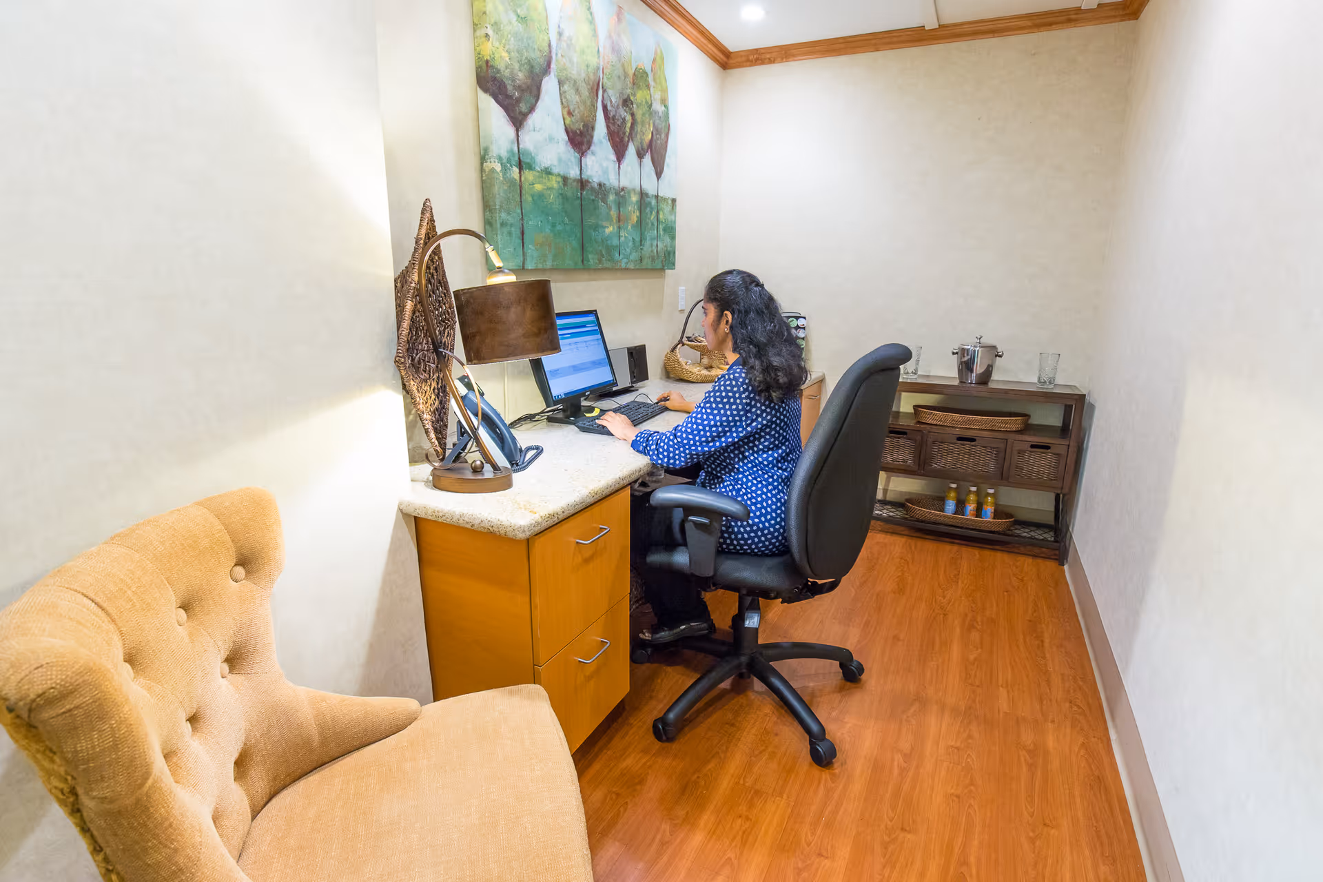 A woman sitting at a desk working on a computer in a small, well-lit office space with wooden flooring. The desk has a lamp, a phone, and some decorative items. There is a cushioned chair nearby and a small shelving unit with baskets and bottles against the wall. A large painting of trees hangs above the desk.