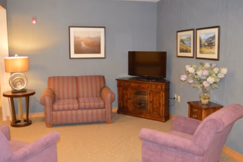 Small living room with a loveseat, two armchairs, a TV on a wooden cabinet, side tables, lamp and framed artwork.
