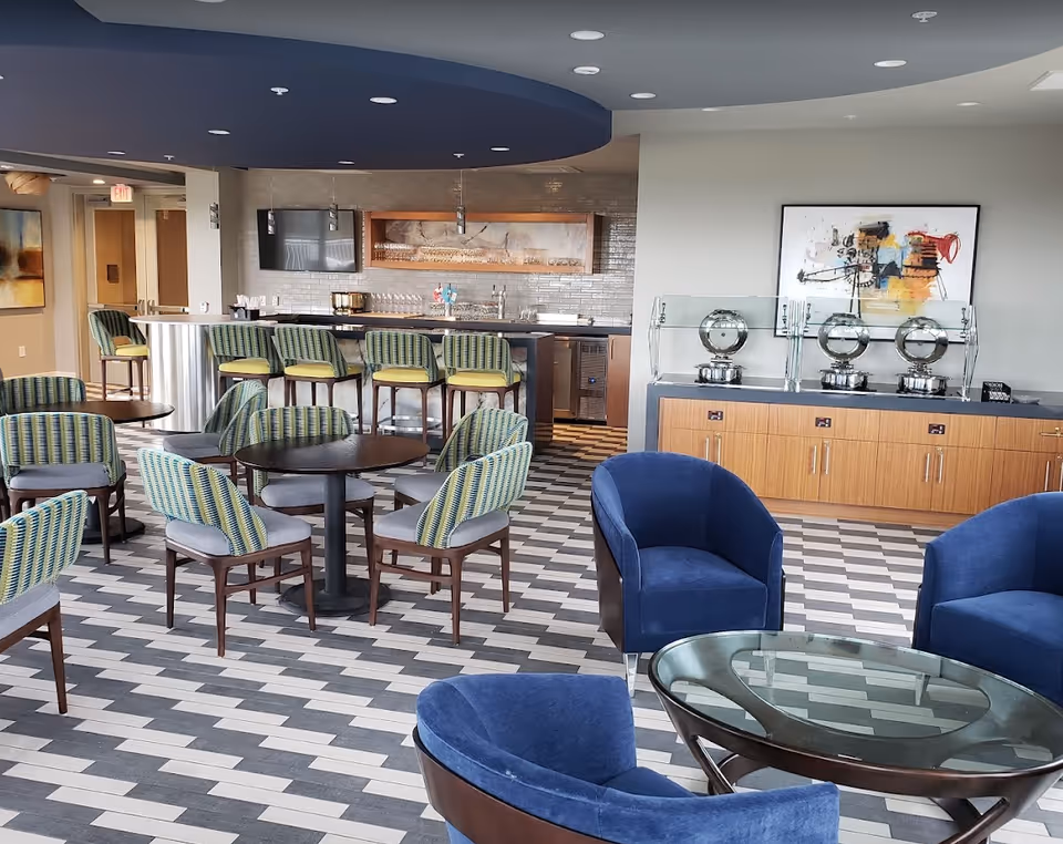 A modern common area in a retirement living facility featuring multiple round tables with green and gray striped chairs, a bar counter with green cushioned bar stools, a sideboard with three covered serving dishes, and blue armchairs around a glass-top table. The floor has a patterned tile design, and there is abstract artwork on the walls.