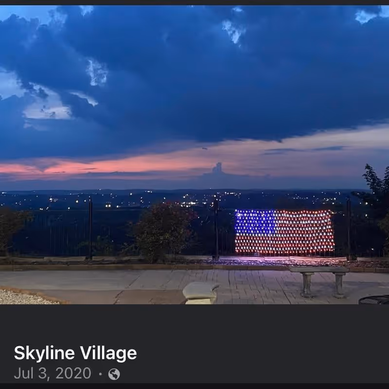 Outdoor evening view with a cloudy sky and a distant cityscape. In the foreground, there is a paved patio area with a bench and a decorative American flag made of lights attached to a fence.
