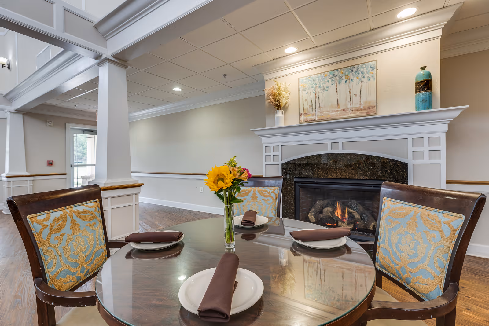 A cozy dining area with a round glass table set for four, each place setting having a white plate and a brown napkin. The chairs have wooden frames with blue and gold patterned cushions. In the background, there is a lit fireplace with a white mantel decorated with a vase of dried flowers, a blue decorative jar, and a painting of trees. The room has wooden flooring and light-colored walls with white trim.