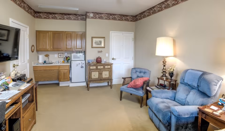 A cozy living area in a senior living facility featuring a blue recliner and a matching armchair with a red pillow. There is a wooden side table with a lamp and decorative items next to the chairs. The room also includes a small kitchenette with wooden cabinets, a mini refrigerator, and a microwave. A flat-screen TV is mounted on the wall opposite the seating area. The walls are beige with a floral border near the ceiling.