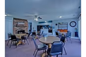 A common area in Prairie Villa featuring multiple round tables with chairs arranged around them. The room has a brick fireplace on one side, a ceiling fan, a clock on the wall, and a bulletin board. The space appears well-lit with natural light coming through windows on the right.