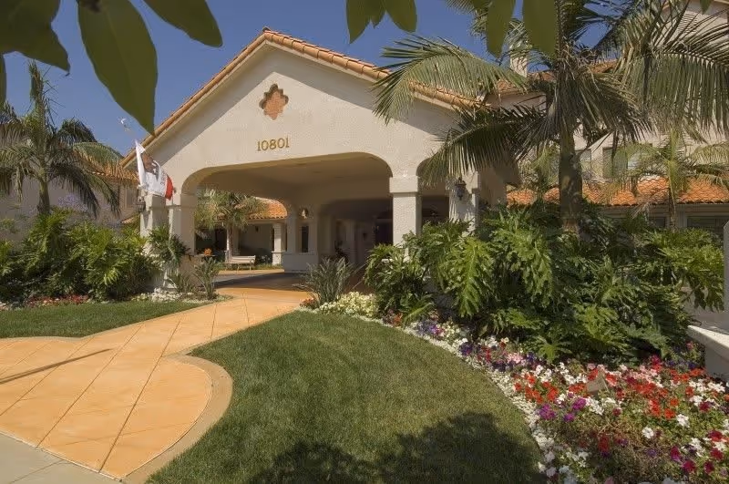 Entrance of a senior living facility with a covered driveway, surrounded by lush green plants, palm trees, and colorful flower beds under a clear blue sky.