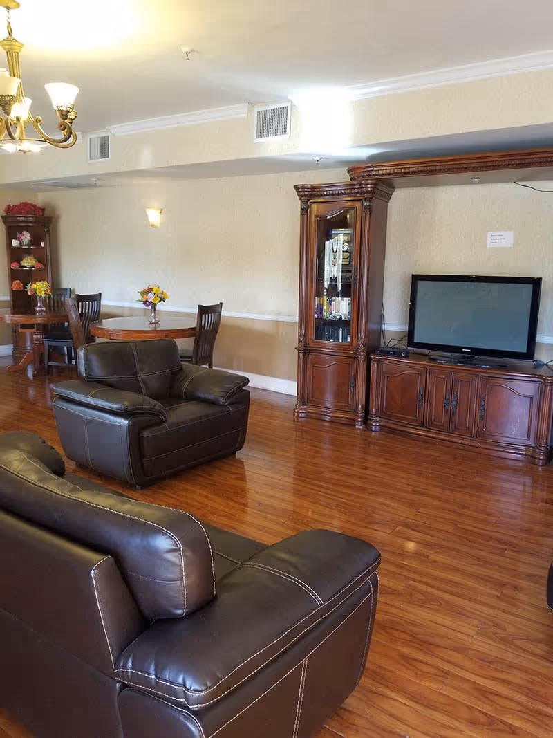 A senior living facility common area with dark brown leather armchairs and a sofa, wooden flooring, a wooden entertainment center with a flat-screen TV, and a round wooden dining table with chairs and flower vases. The room is softly lit with a chandelier and wall sconces.