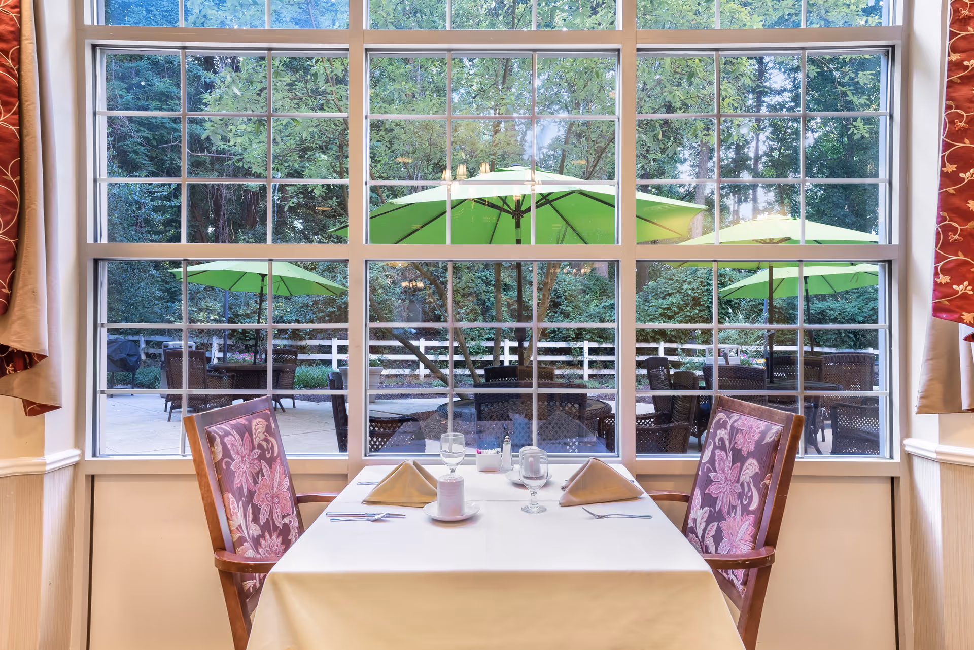 A dining table set for two with folded napkins, glasses, and condiments, positioned in front of a large window. Outside the window, there are green patio umbrellas, outdoor tables, chairs, and lush greenery with trees and a white fence.