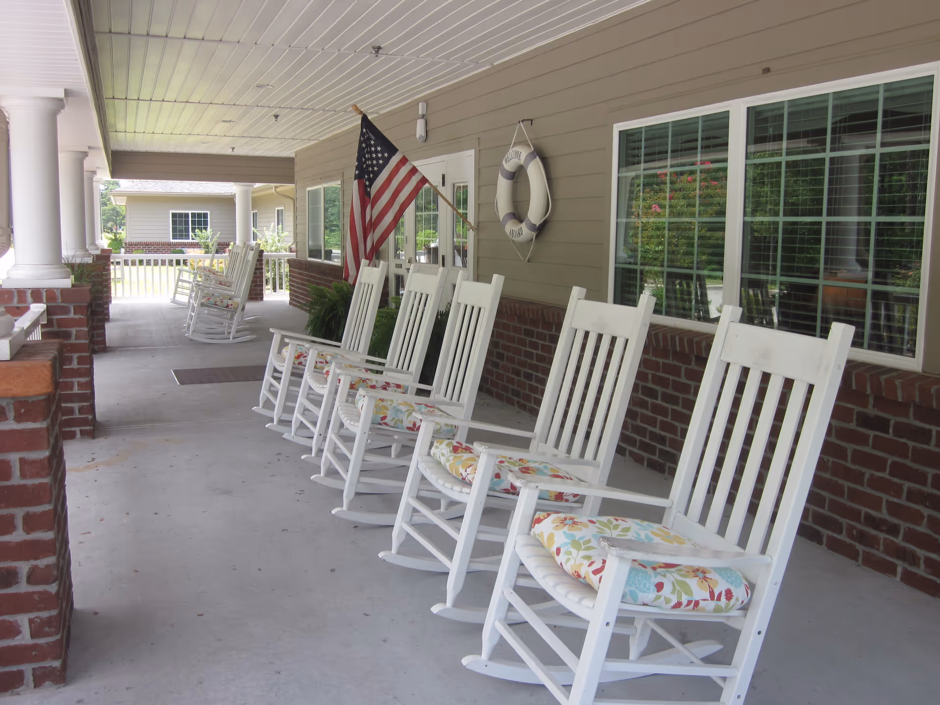 Covered porch area with a row of white wooden rocking chairs with floral cushions. An American flag is mounted on the wall next to a life preserver decoration that says 'Welcome Aboard'. The porch has brick half-walls and white columns, with windows along the building wall.