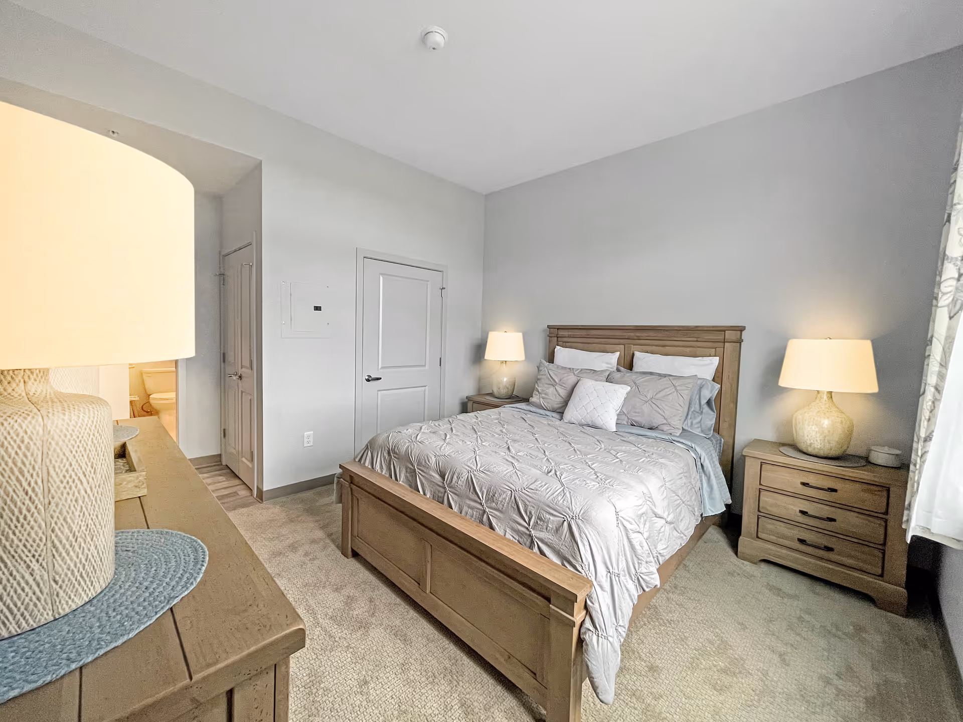 A bedroom with a wooden bed frame and a neatly made bed with gray bedding and multiple pillows. There are two matching wooden nightstands on either side of the bed, each with a lamp. A dresser with a lamp is visible in the foreground on the left. The room has light gray walls, beige carpet, and a window with patterned curtains on the right. A door and a partially visible bathroom are seen in the background.