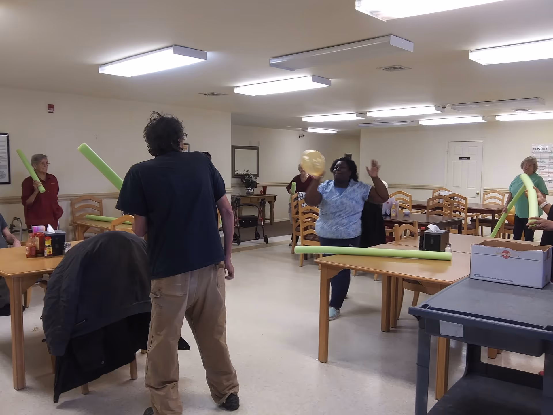 A group of people in a communal dining/activity room playing with pool noodles and a balloon around tables and chairs.