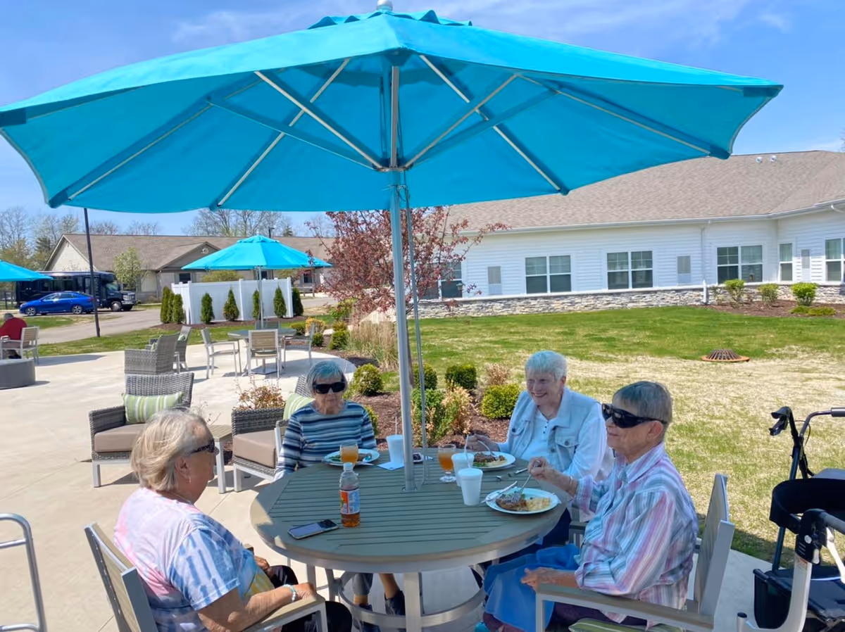 Four elderly women sitting around a round outdoor table under a large blue umbrella, eating a meal and enjoying a sunny day at a senior living facility. The background shows a well-maintained lawn, shrubs, and a white building with multiple windows.