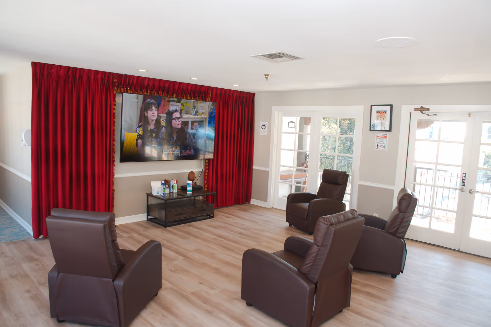 A cozy living room area in an assisted living facility with four brown recliner chairs arranged facing a large flat-screen TV mounted on the wall. The TV is framed by red curtains. Below the TV is a small table holding various items. The room has light wood flooring and white walls with a chair rail. There are glass double doors letting in natural light on the right side of the room.