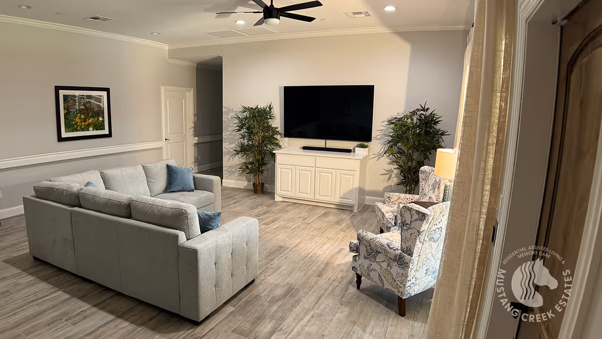 A cozy living room with a large gray sectional sofa adorned with blue pillows, two patterned armchairs, a white TV console with a flat-screen TV mounted above it, two tall green plants flanking the TV, a ceiling fan, and a framed floral picture on the wall. The room has wood-look flooring and neutral-colored walls.