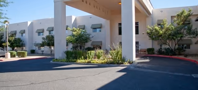 Entrance and porte-cochere of a two-story beige assisted living building with green awnings, landscaping, and a driveway.