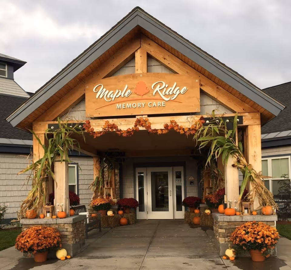 Entrance of Maple Ridge Memory Care facility decorated with autumn-themed items including pumpkins, corn stalks, orange flowers, and a garland of fall leaves hanging under a wooden sign with the facility's name.