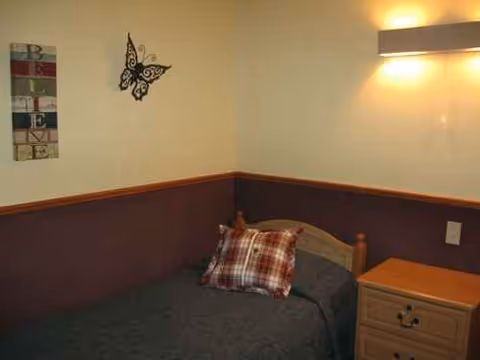 Small bedroom with a single bed, plaid pillow, nightstand, and soft lighting.