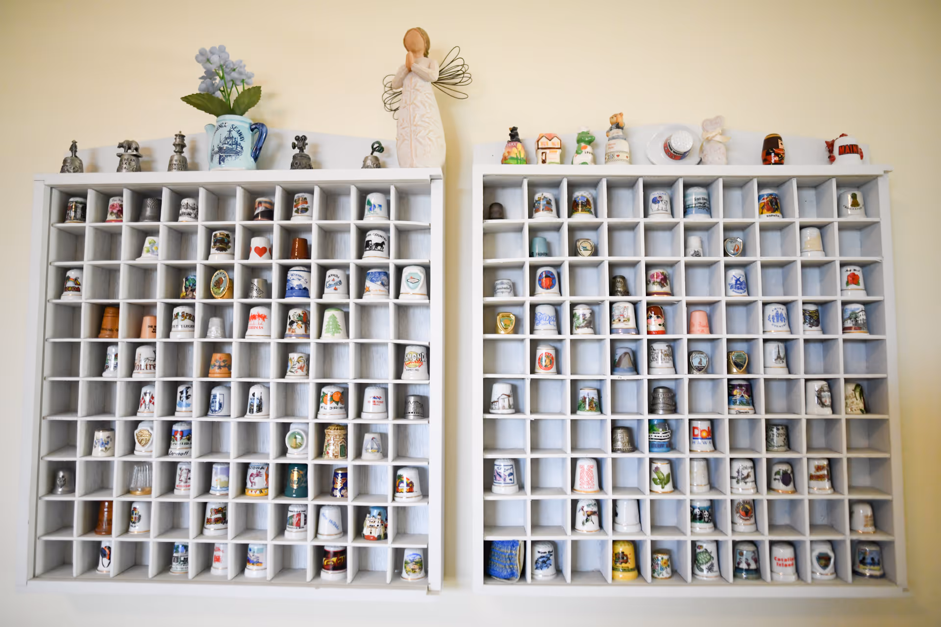 Two white wooden display shelves mounted on a beige wall, each with multiple small compartments holding a collection of decorative thimbles. On top of the left shelf is a small blue vase with purple flowers and a figurine of an angel with wire wings. The right shelf has various small decorative items on top.