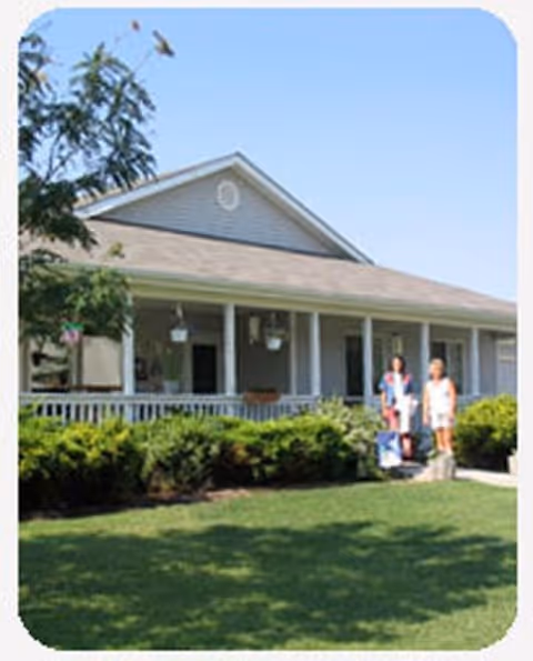 A single-story building with a wide porch and white railing, surrounded by green bushes and a well-maintained lawn. Two people stand on the porch steps under a clear blue sky.