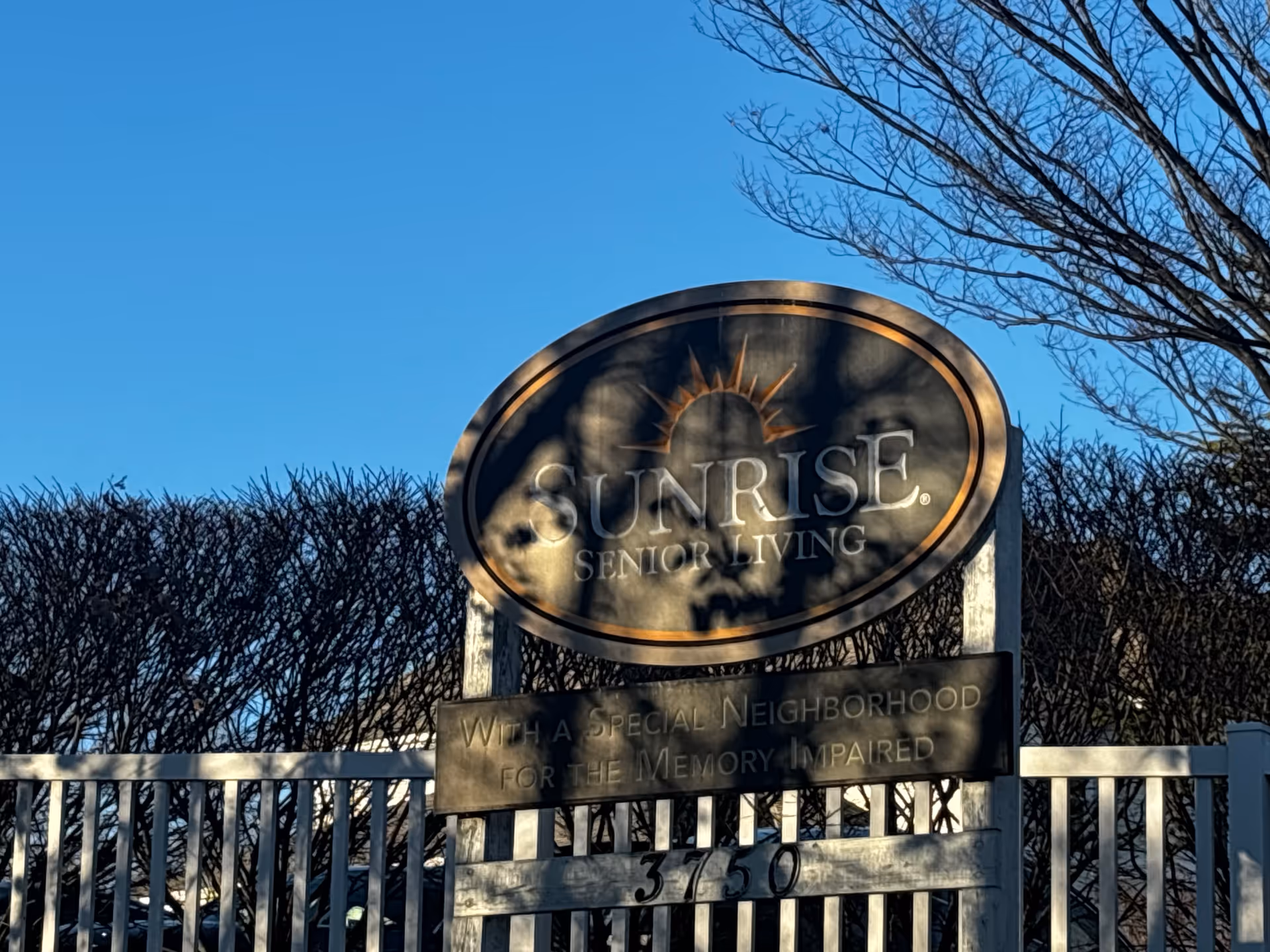 Outdoor sign for Sunrise Senior Living with a special neighborhood for the memory impaired, set against a clear blue sky and leafless trees and bushes.