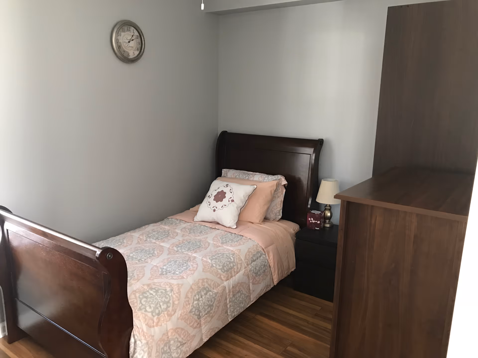 Small bedroom with a wooden single sleigh bed, patterned bedding, a nightstand with lamp, and a tall wooden dresser.