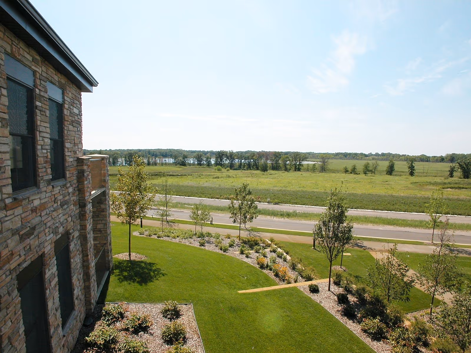 View from a building showing a landscaped garden with green grass, small trees, and flower beds. A road runs beyond the garden, and there is a wide open field with trees and a body of water in the distance under a partly cloudy sky.