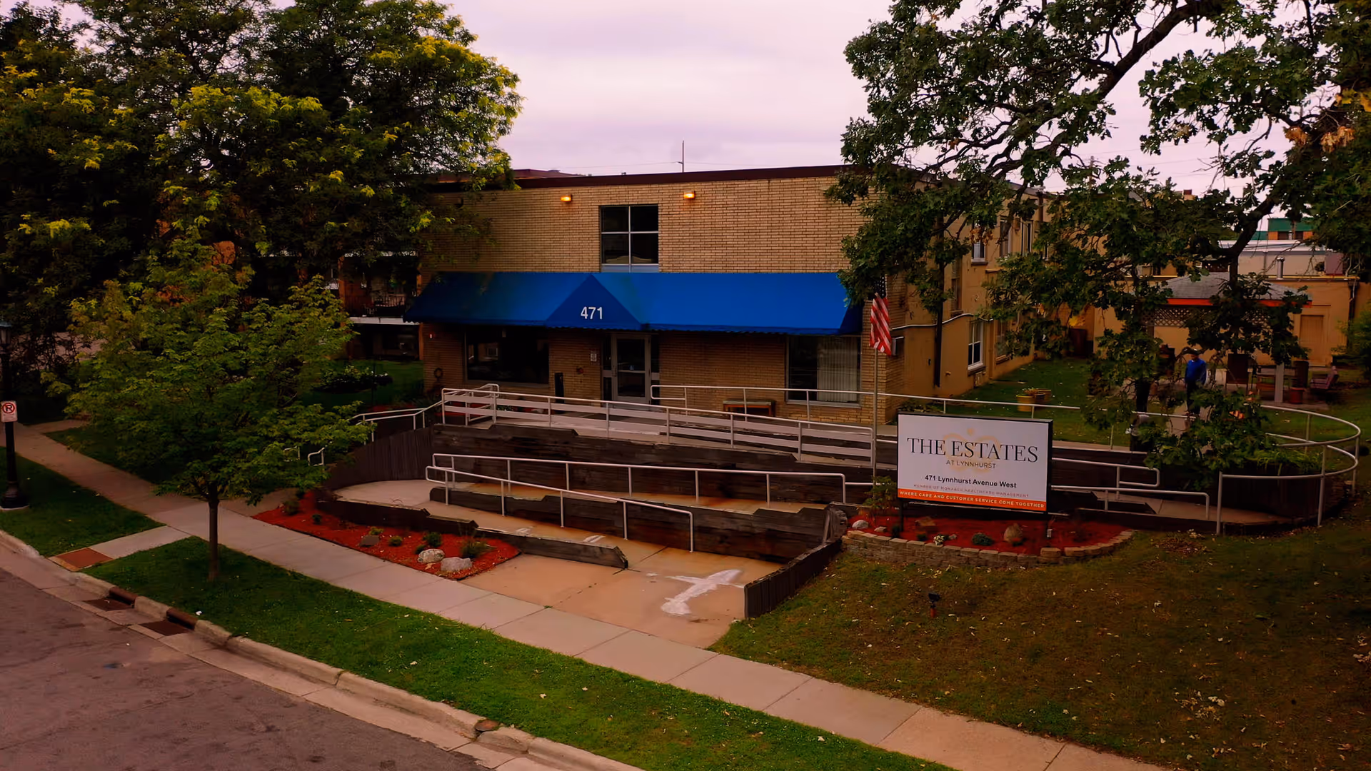 Exterior view of The Estates at Lynnhurst building with a blue awning over the entrance marked 471. There is a wheelchair accessible ramp leading to the entrance, surrounded by trees and landscaping with red mulch and rocks. A sign in front of the building displays the facility name and address.