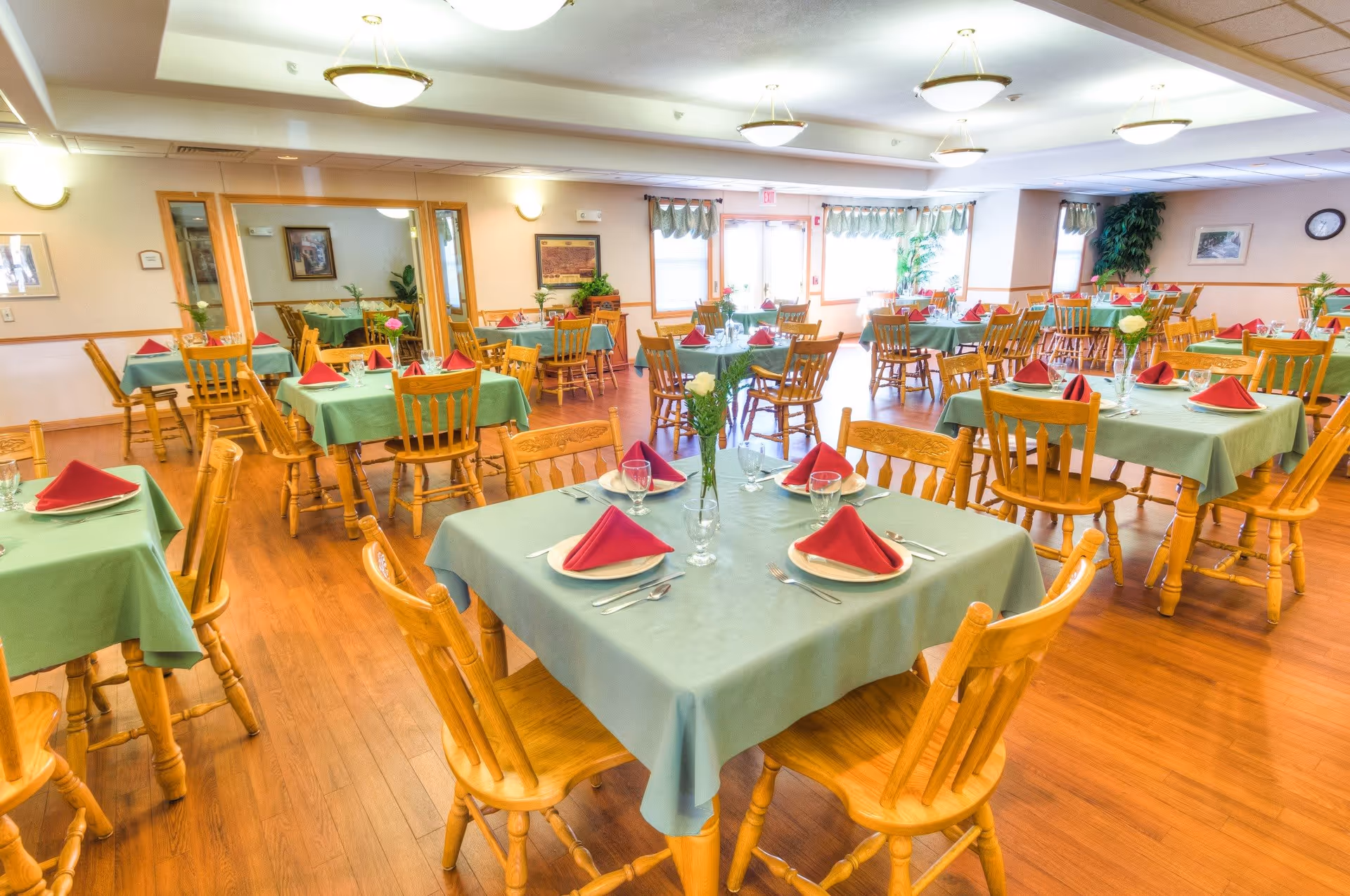A bright dining room with multiple wooden tables and chairs arranged neatly. Each table is covered with a green tablecloth and set with white plates, silverware, glasses, and red folded napkins. There are small flower vases with white flowers on the tables. The room has wooden flooring, light-colored walls, and large windows letting in natural light. Ceiling lights and wall sconces illuminate the space.