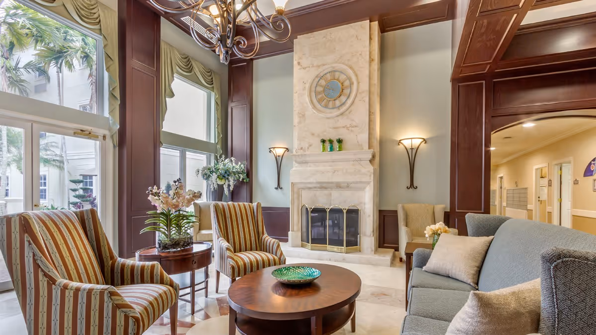 Sunlit lounge with striped armchairs, a sofa and round wooden coffee table facing a marble fireplace topped by a large clock.