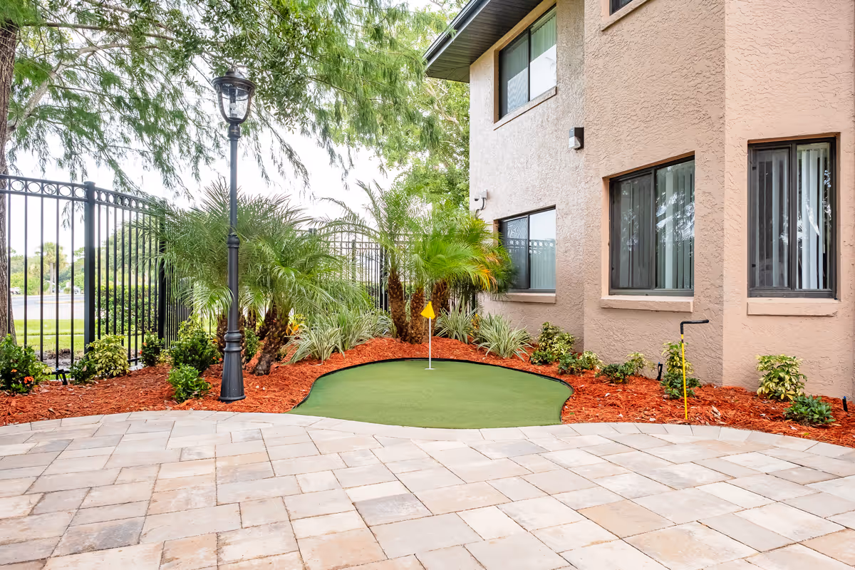 Small outdoor courtyard with a putting green, lamppost, palm plants and the exterior wall of a beige building.