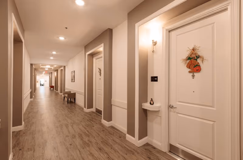 Long interior hallway in a senior living facility with wood-look floors, recessed lighting, seating, and decorated apartment doors.