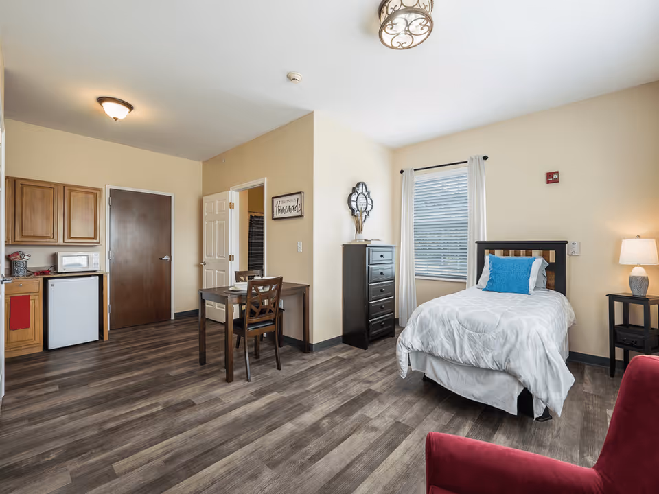 A bright and clean senior living studio apartment featuring a single bed with white bedding and a blue pillow, a black chest of drawers, a small dining table with two chairs, a kitchenette with wooden cabinets, a mini fridge, and a microwave. The room has wood-look flooring, beige walls, a window with white curtains, and a red armchair in the foreground.