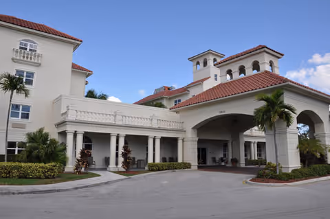 Exterior view of a senior living facility with Mediterranean-style architecture, featuring beige walls, red tile roofs, columns, and a covered entrance driveway. Palm trees and landscaping surround the building under a clear blue sky.