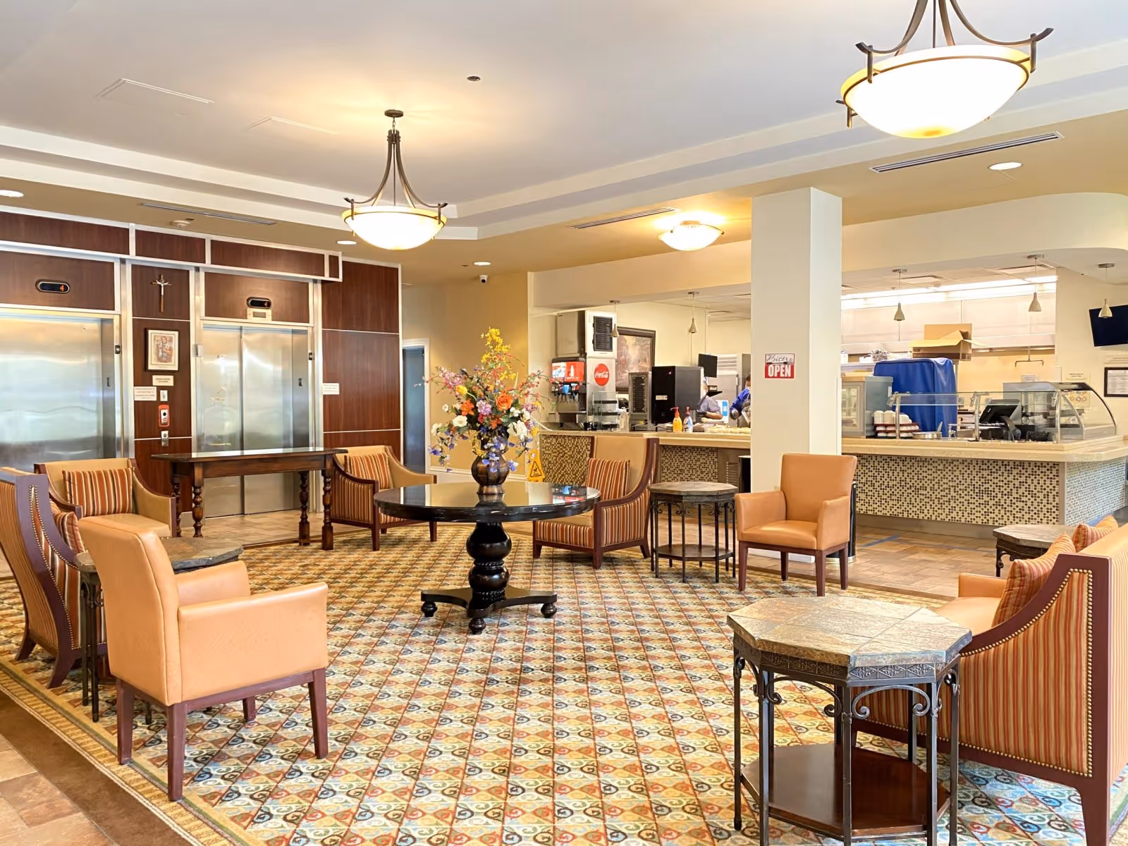 A spacious common area in a senior living facility featuring a patterned carpet, multiple armchairs and small tables arranged around a central round table with a floral arrangement. In the background, there is a counter area with kitchen equipment and a sign that says 'OPEN'. Two elevators with wood paneling are visible on the left side of the image.