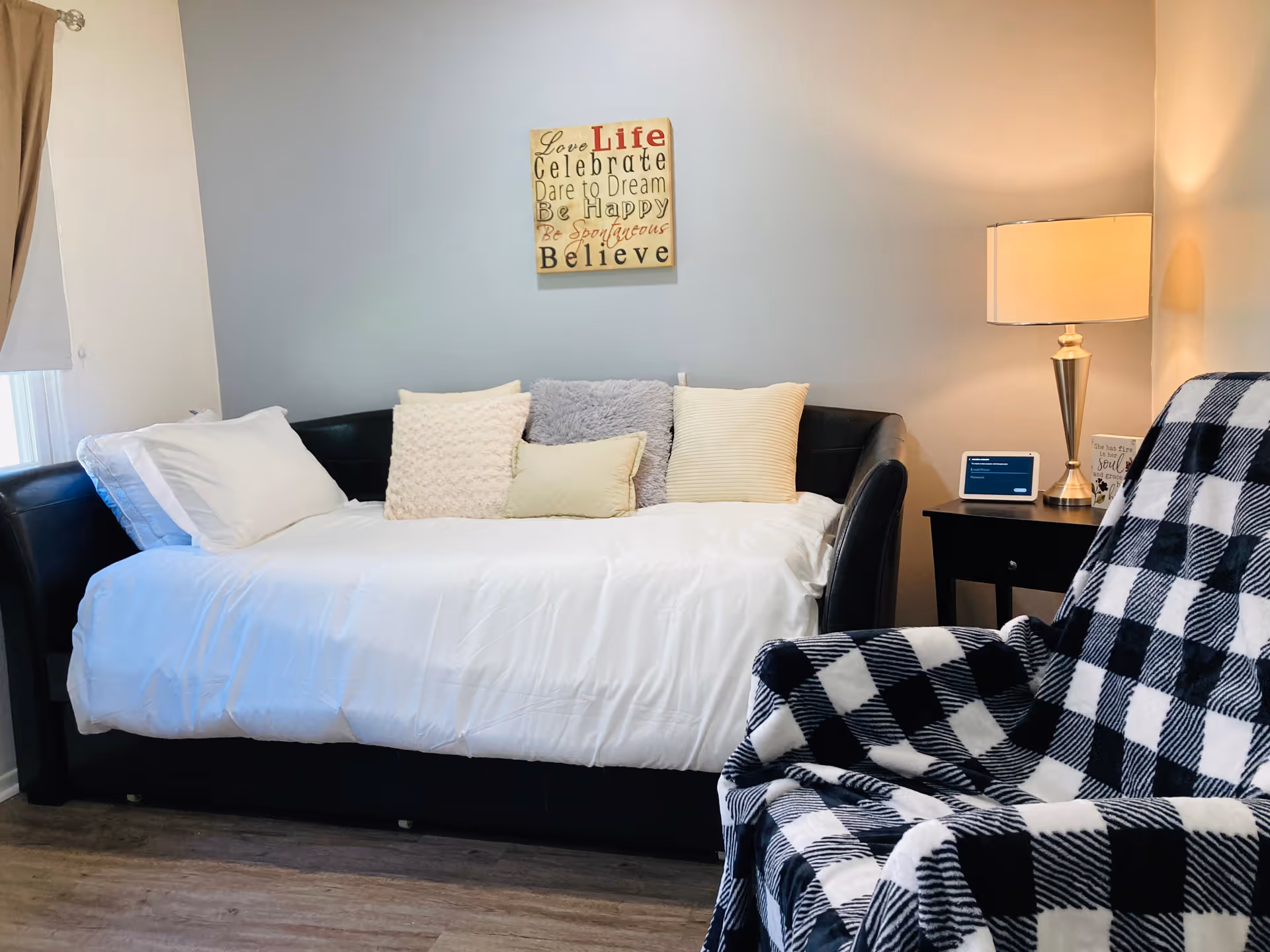 A cozy room with a black daybed covered with white bedding and multiple pillows against a light blue wall. Above the bed is a decorative wall art with inspirational words. To the right, there is a black side table with a lamp and a small digital device. In the foreground, a chair is draped with a black and white checkered blanket.