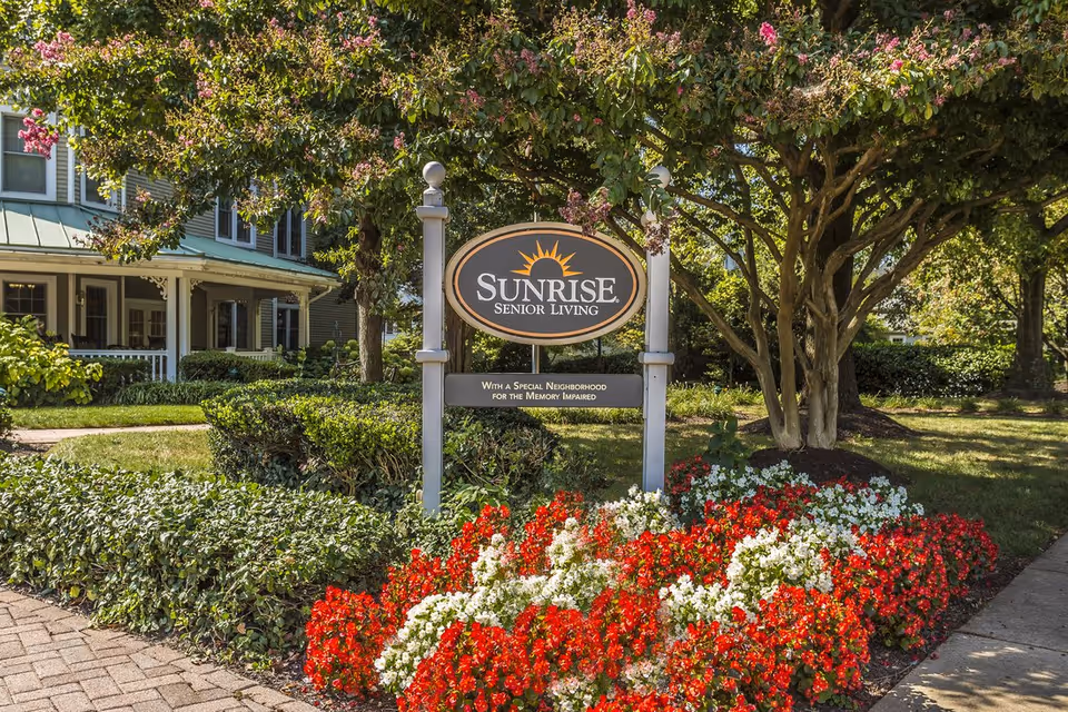 An entrance sign reading "Sunrise Senior Living" surrounded by flowering beds, trees, and a porch-front building.