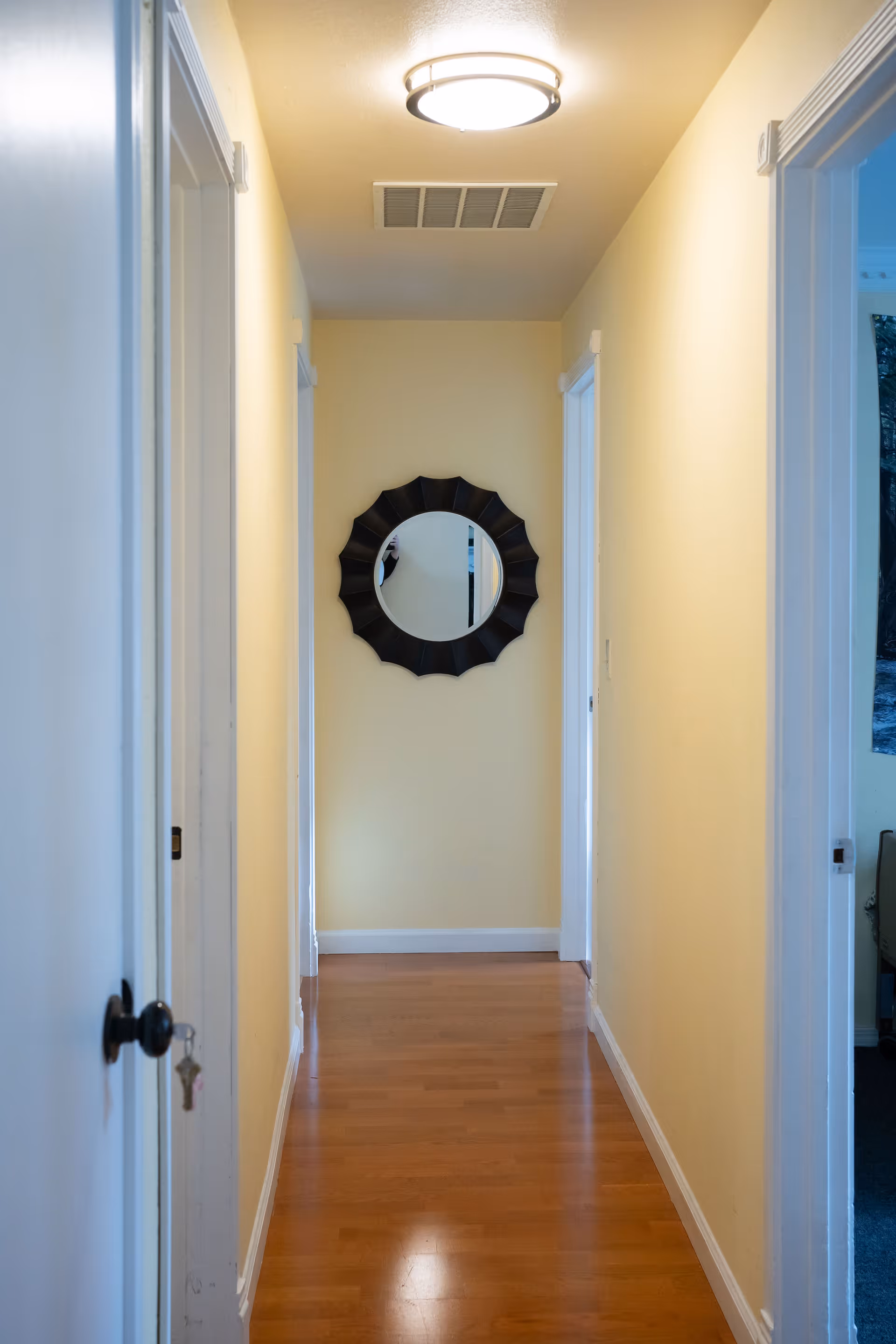 A narrow, well-lit hallway with yellow walls, hardwood floors, a ceiling light, and a decorative round mirror at the end.