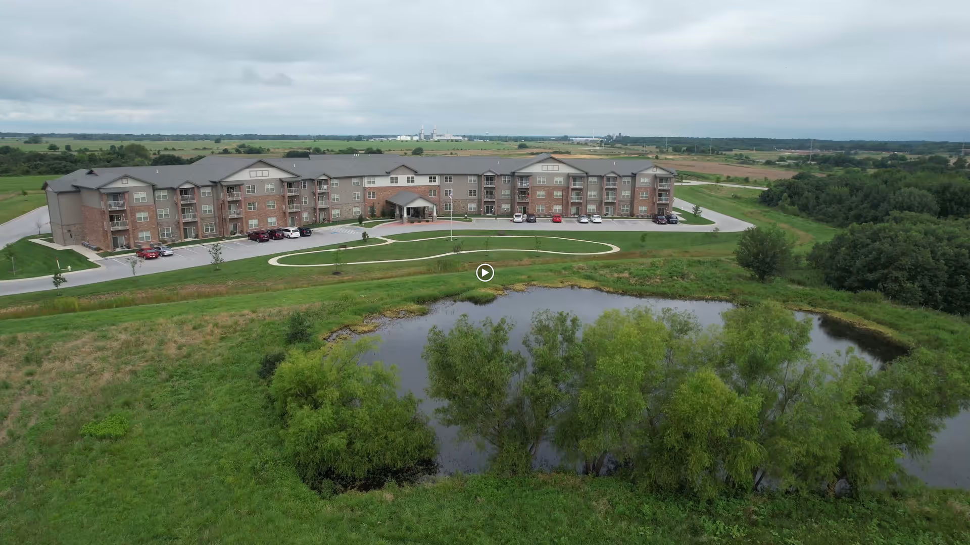 Aerial view of The Wildwood Senior Living facility showing a large three-story building with multiple windows and a covered entrance. The building is surrounded by green lawns, a small pond with trees, and open fields under a cloudy sky.