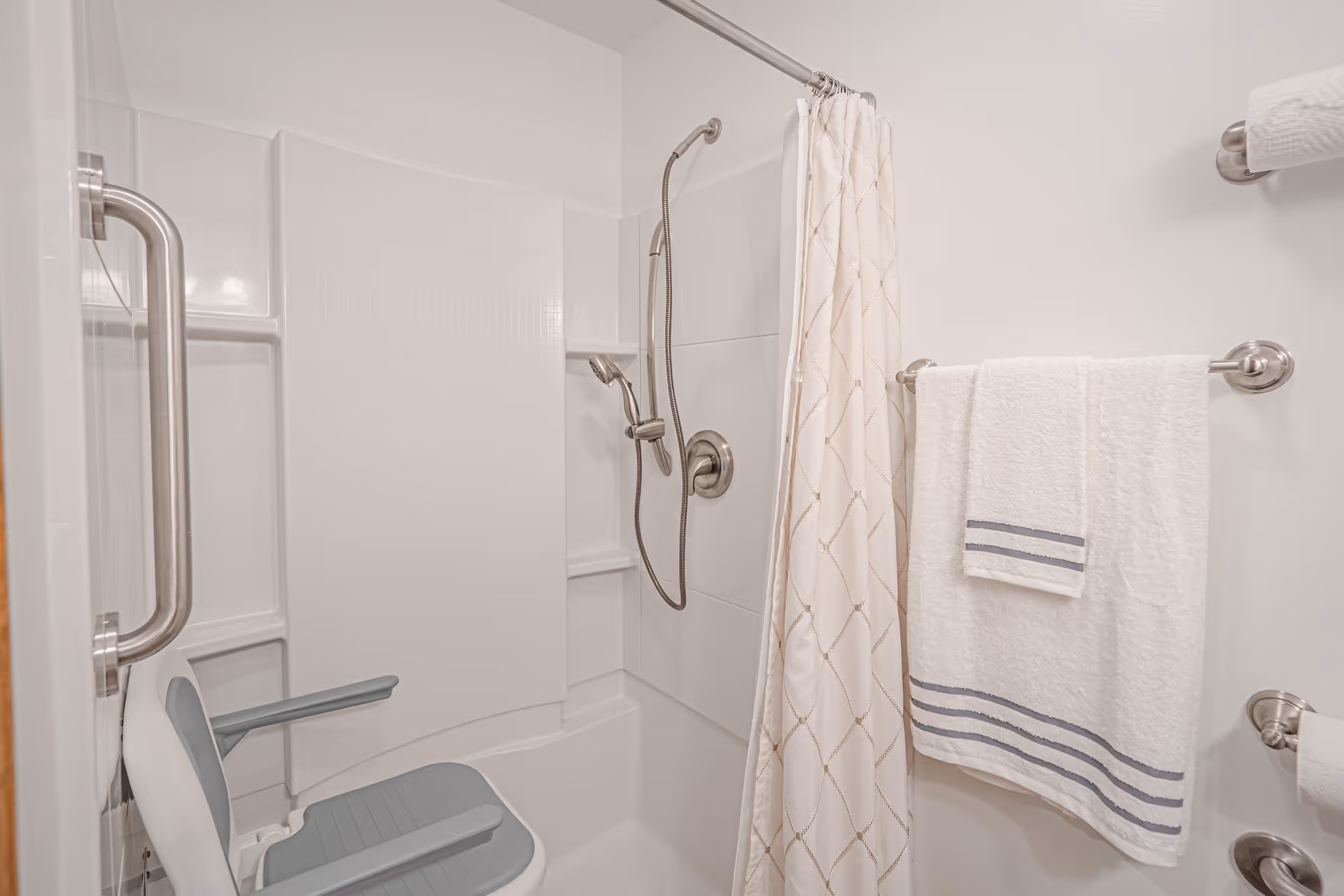 A bathroom shower area with a white bathtub and a handheld showerhead mounted on the wall. There is a white shower curtain with a subtle pattern, a white towel with blue stripes hanging on a towel rack, and a shower chair with armrests inside the tub. A grab bar is installed on the wall for safety.
