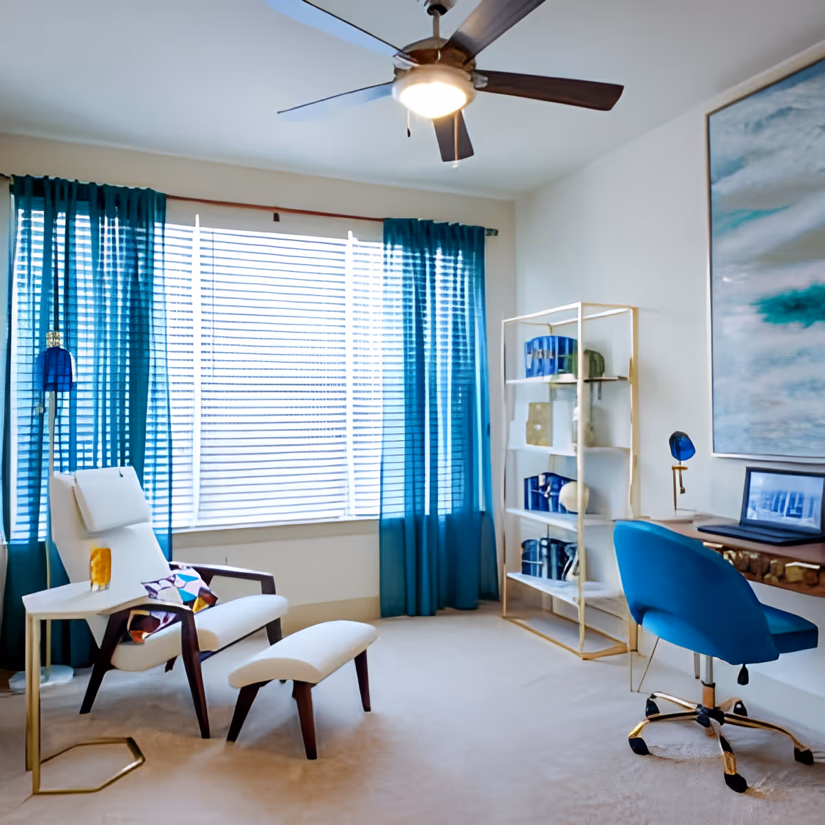 A bright room with a white cushioned armchair and matching ottoman, a small side table with a glass of orange drink, teal curtains covering a large window with blinds, a ceiling fan with light, a gold-framed shelving unit with decorative items, a wooden desk with a blue swivel chair, a blue desk lamp, and a large abstract painting on the wall.