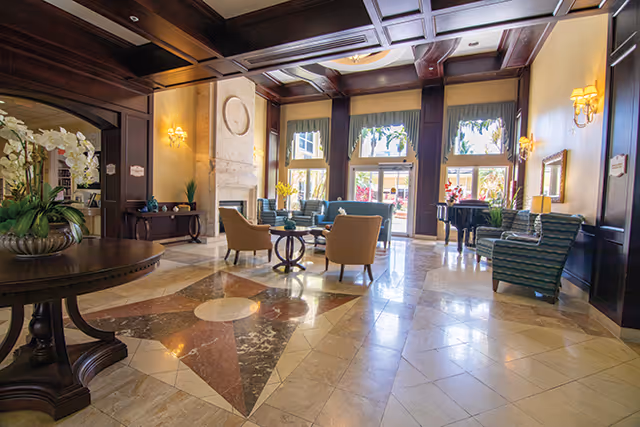 Spacious and elegant common area with polished marble floors featuring a star pattern, comfortable armchairs, a grand piano near large windows, and warm lighting from wall sconces and ceiling fixtures.