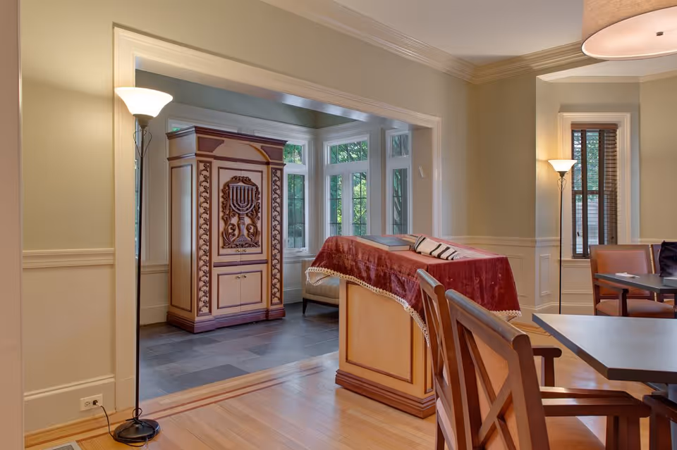 Interior view of a room with wooden flooring and beige walls featuring a large wooden cabinet with a menorah design, a lectern covered with a red velvet cloth, wooden chairs, tables, and two standing lamps near windows with white trim.