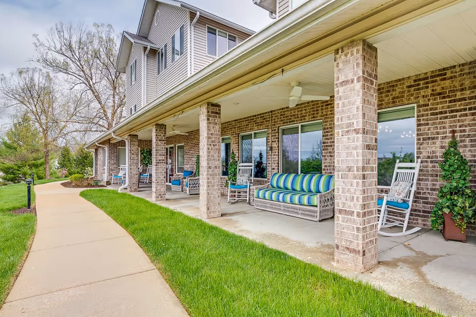 Covered outdoor patio area of a senior living facility with brick pillars, rocking chairs, a striped cushioned bench, potted plants, and a concrete walkway alongside a green lawn.